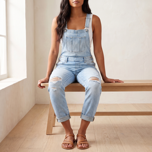 Bianca denim overalls in Light Blue worn seated on an oak bench, showing the rolled hems