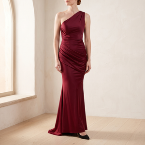 Cecily one shoulder satin maxi dress in Burgundy by a window in a bright room with plaster walls