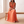 Cristina pleated maxi dress in Orange worn seated on a pale oak bench