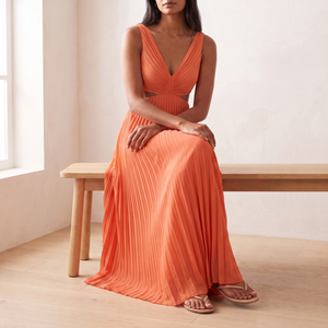 Cristina pleated maxi dress in Orange worn seated on a pale oak bench