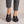 Elinor round toe flats in Black worn with cropped gray trousers on a light gray studio backdrop
