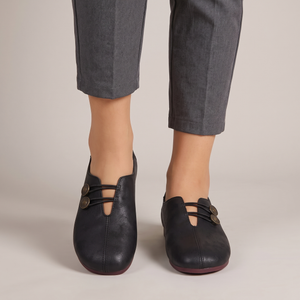 Elinor round toe flats in Black worn with cropped gray trousers on a light gray studio backdrop