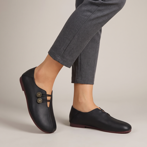 Elinor round toe flats in Smooth Black on a woman's feet with cropped gray trousers, light gray studio backdrop