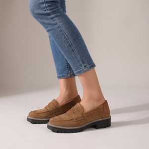 Gloria suede penny loafers in Brown worn with blue jeans on a light gray studio backdrop