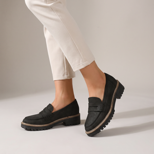 Gloria suede penny loafers in Black worn with ecru cropped trousers on a light gray studio backdrop