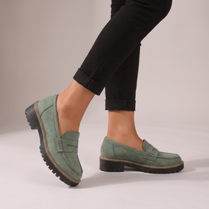 Gloria suede penny loafers in Sage worn with black cropped trousers on a light gray studio backdrop