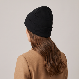 Hattie knit beanie in Black worn with a camel coat, seen from behind on a light gray studio backdrop