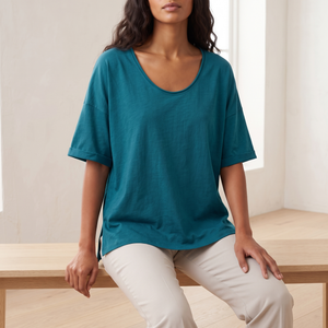 Jemima loose scoop neck blouse in Teal worn seated on an oak bench