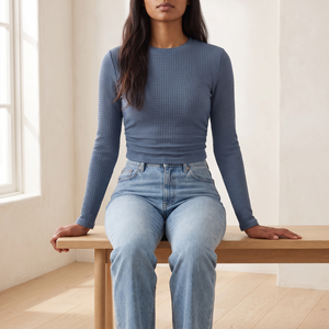 Maribel long sleeve crop top in Blue, seated on an oak bench in a bright room