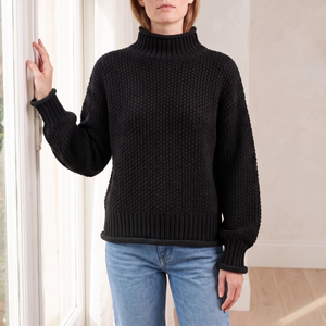 Romane chunky knit turtleneck sweater in Black worn by a tall window