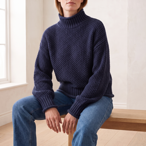 Romane chunky knit turtleneck sweater in Navy Blue worn seated on an oak bench