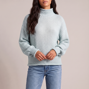 Romane chunky knit turtleneck sweater in Light Blue with jeans on a light gray studio backdrop