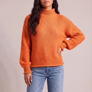 Romane chunky knit turtleneck sweater in Orange with jeans on a light gray studio backdrop