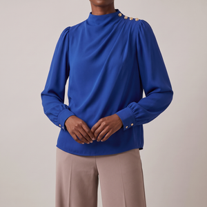 Solange draped mock neck blouse in Blue on a light gray studio backdrop