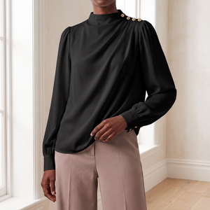 Solange draped mock neck blouse in Black, worn with taupe trousers by a window in a bright room
